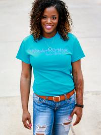 A woman with curly hair, wearing a teal t-shirt and ripped jeans, stands outdoors on a concrete surface, smiling at the camera.