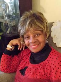 Smiling older woman with short, curly hair sits on a couch, wearing a red patterned top over a black turtleneck, with a watch on her wrist and her hand resting by her face.