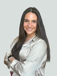 A woman with long dark hair, wearing a light-colored satin blouse and a smartwatch, stands smiling with her arms crossed against a plain light gray background.