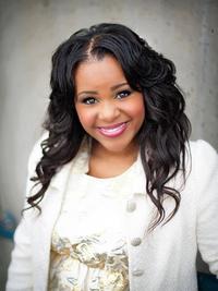 A woman with long, wavy dark hair smiles at the camera. She is wearing a white textured jacket over a light-colored top and stands against a neutral background.