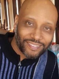 A man with a trimmed beard and a bald head smiles at the camera. He is wearing a dark zip-up sweater with light and dark vertical stripes in the background.