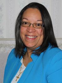 A smiling woman with straight dark hair and glasses wears a bright blue blazer over a white top and a gold cross necklace, standing in front of a patterned wallpaper background.