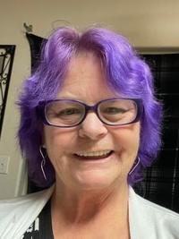 A woman with short, bright purple hair and glasses smiles at the camera. She is wearing dangling earrings and a light-colored top. The background shows part of a room with a black curtain.