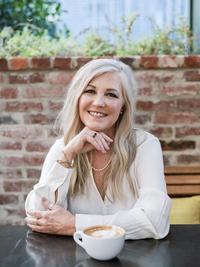 A smiling woman with long blonde hair, wearing a light blouse and necklace, sits at a table with a cup of coffee in front of her. Brick wall and greenery are visible in the background.