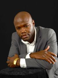 A man in a gray suit and white shirt leans forward with his arms crossed on a black surface, wearing a gold watch, against a plain black background.