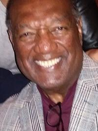 A smiling older man in a checked blazer and maroon shirt poses for a close-up photo. He has short hair and is indoors, with part of another person visible beside him.