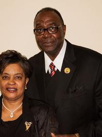 A man and a woman dressed in formal black attire stand together, both smiling. The man wears glasses, a black suit, and a striped tie; the woman wears a black jacket with a brooch and a necklace, against a plain background.
