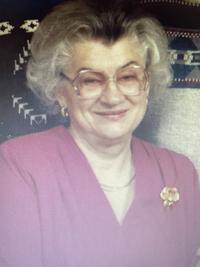 An older woman with gray hair and glasses smiles while wearing a red top with a flower brooch, sitting in front of a patterned background.