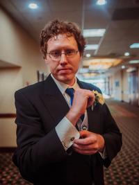 A man in a suit adjusts his cufflinks while standing in a hallway with patterned carpet and soft overhead lighting. He has glasses, curly brown hair, and a boutonnière on his lapel.