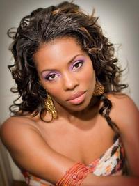 A woman with long, curly hair and gold earrings poses for the camera. She wears a strapless patterned top, bold purple eye makeup, and orange bracelets, looking directly at the viewer with a soft, serious expression.
