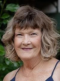 A smiling older woman with wavy, shoulder-length light brown hair stands outdoors, wearing a dark sleeveless top. Green foliage is visible in the background.