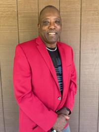 A man stands in front of a wooden wall, smiling. He is wearing a bright red blazer over a black shirt and a silver necklace. His hands are clasped in front of him.