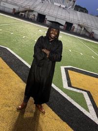 A person wearing a black graduation gown and cap stands smiling on a football field, with bleachers in the background and arms crossed in front.