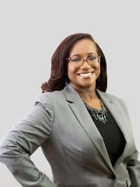A woman wearing glasses, a gray blazer, and a black top smiles confidently at the camera against a plain, light gray background.