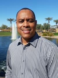 A man in a striped shirt stands smiling in front of a pond with a fountain, palm trees, and a clear blue sky in the background.