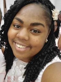 A young woman with braided hair adorned with a light-colored flower accessory smiles at the camera. She is wearing a white top and is indoors, with a blurred background.