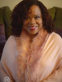 A woman with curly hair smiles at the camera, wearing a soft pink shawl with a fur trim, seated indoors against a green couch.
