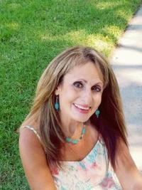 A woman with long light brown hair smiles outdoors, wearing a floral dress and turquoise jewelry. She is sitting on grass near a sunlit sidewalk.