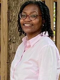 A woman wearing glasses and a light pink button-up shirt smiles while standing outdoors in front of a wooden fence and tree.