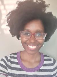 Smiling person with natural curly hair, wearing glasses and a striped shirt with purple trim, standing in front of a neutral-colored wall.