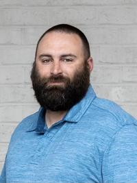 A man with a full dark beard and short hair wearing a light blue collared shirt stands in front of a light-colored brick wall, looking at the camera with a neutral expression.