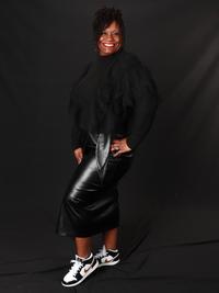 A woman stands smiling against a black background, wearing a black sweater, a shiny black leather skirt, and black-and-white sneakers. She has curly hair styled to one side and poses with one hand on her hip.