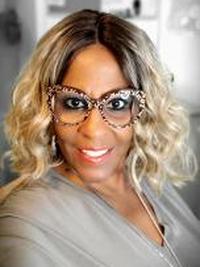 A woman with shoulder-length blonde curly hair, wearing large, patterned eyeglasses and a light gray top, smiles at the camera against a softly blurred background.