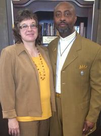 A woman and a man stand side by side in front of a bookshelf. Both are wearing brown suits, and the woman has glasses and curly hair. The man has a beard and is wearing a white shirt under his suit.