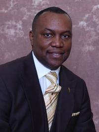 A man wearing a dark suit, white shirt, and a gold patterned tie, smiling while seated in front of a brown textured background.