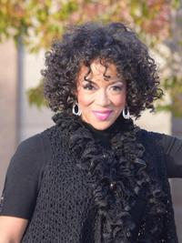 A woman with curly dark hair and hoop earrings smiles outdoors. She is wearing a black textured scarf and a black top, with blurred autumn leaves in the background.