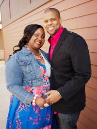 A smiling couple stands close together in front of a brown wall. The woman wears a blue floral dress and denim jacket, while the man wears a black jacket and a bright pink shirt. They are holding hands and posing for the photo.
