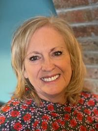 A woman with blonde hair smiles at the camera. She is wearing a floral top with a red and black pattern. Behind her is a teal wall and a section of exposed brick.