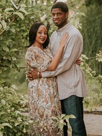 A couple stands closely together outdoors, surrounded by greenery. The woman wears a patterned dress and the man wears a light button-down shirt and jeans. They smile gently, with the woman’s hand resting on the man’s chest.