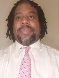 A man with short dreadlocks and a goatee is wearing a white shirt and a light pink striped tie, standing in front of a plain beige background.