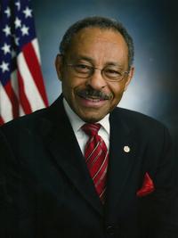 A man wearing glasses, a dark suit, red striped tie, and red pocket square smiles in front of a blurred U.S. flag and a dark blue background.