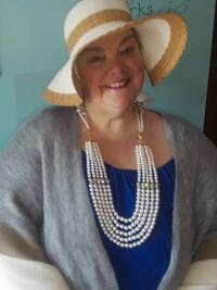 A smiling woman wearing a wide-brimmed straw hat, a large layered pearl necklace, a blue top, and a gray shawl stands in front of a light blue background.