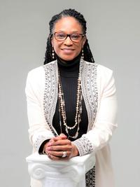 A woman with long braided hair, glasses, and layered necklaces wears a black turtleneck and a light-colored, embroidered jacket. She is smiling and leaning on a white decorative pedestal against a plain background.