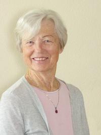 An older woman with short white hair smiles at the camera. She is wearing a light pink top, a gray cardigan, and a necklace with a small red pendant, standing against a plain beige background.