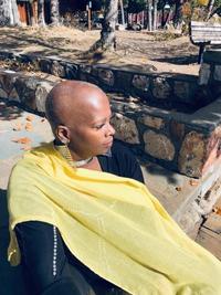 A person with a shaved head sits outdoors on a sunny day, draped in a yellow scarf over a black top, looking to the side. Stone benches and scattered leaves are visible in the background.