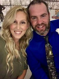 A smiling woman with long blonde hair and a man with a beard wearing a blue shirt and floral tie sit close together at an indoor event with string lights.