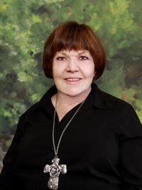 A woman with short brown hair and a black shirt, wearing a large silver cross necklace, stands in front of a green, leafy backdrop.