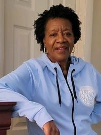 An older woman with short curly hair, wearing a light blue zip-up hoodie, stands indoors with her arm resting on a wooden surface, smiling softly at the camera. A white door is visible in the background.