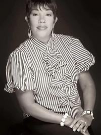 A woman poses for a formal portrait, wearing a striped blouse with ruffles, earrings, and bracelets. She is seated, facing the camera, with a neutral expression against a dark background. The photo is in black and white.