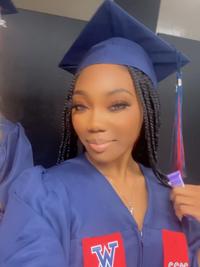 A young woman wearing a blue graduation cap and gown with long braids smiles at the camera. She holds the tassel of her cap and stands indoors, appearing happy and proud.